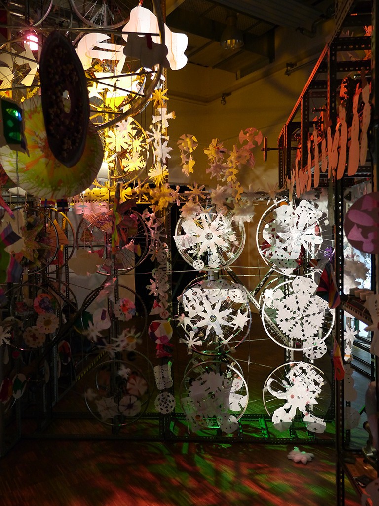 Objekt in der Lichtmaschinerie Mitmach-Ausstellung: beleuchtete Konstruktion mit zahlreichen Rädern, alles mit Schneeflocken und Eiskristallen aus Papier bestückt.